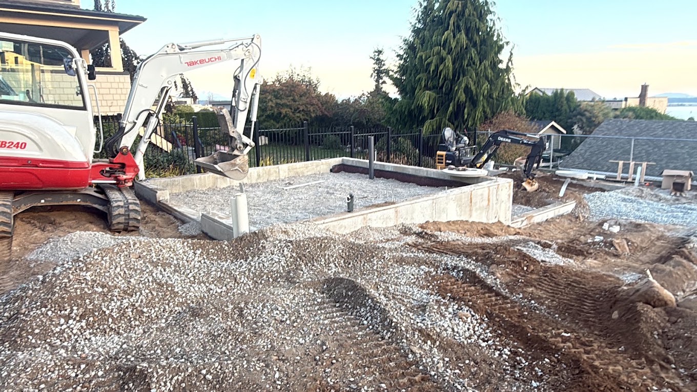 Excavators beside a prepared foundation with gravel base and underground service pipes